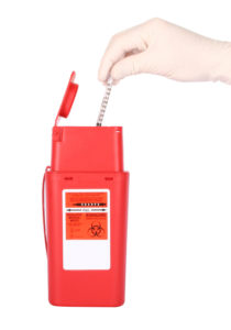 A hand putting a syringe into a sharps medical waste container.